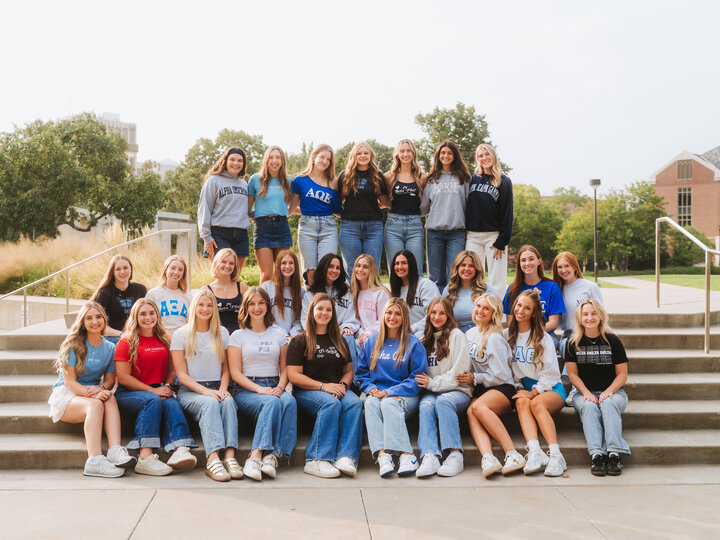 Group of women seated and standing on outdoor steps, dressed casually, with campus buildings in the background.